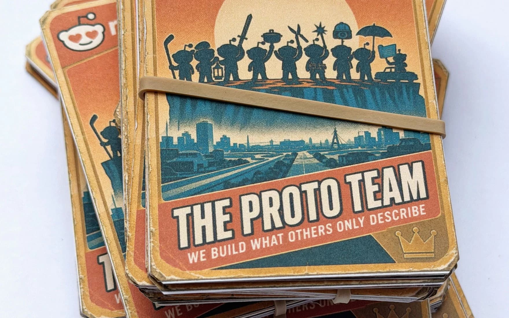 A stack of the Proto team's cards held by a rubber band. The top card shows the team as silhouettes on a cliff against a setting sun, each holding a tool, above a city, and reads "The Proto Team: we build what others only describe".