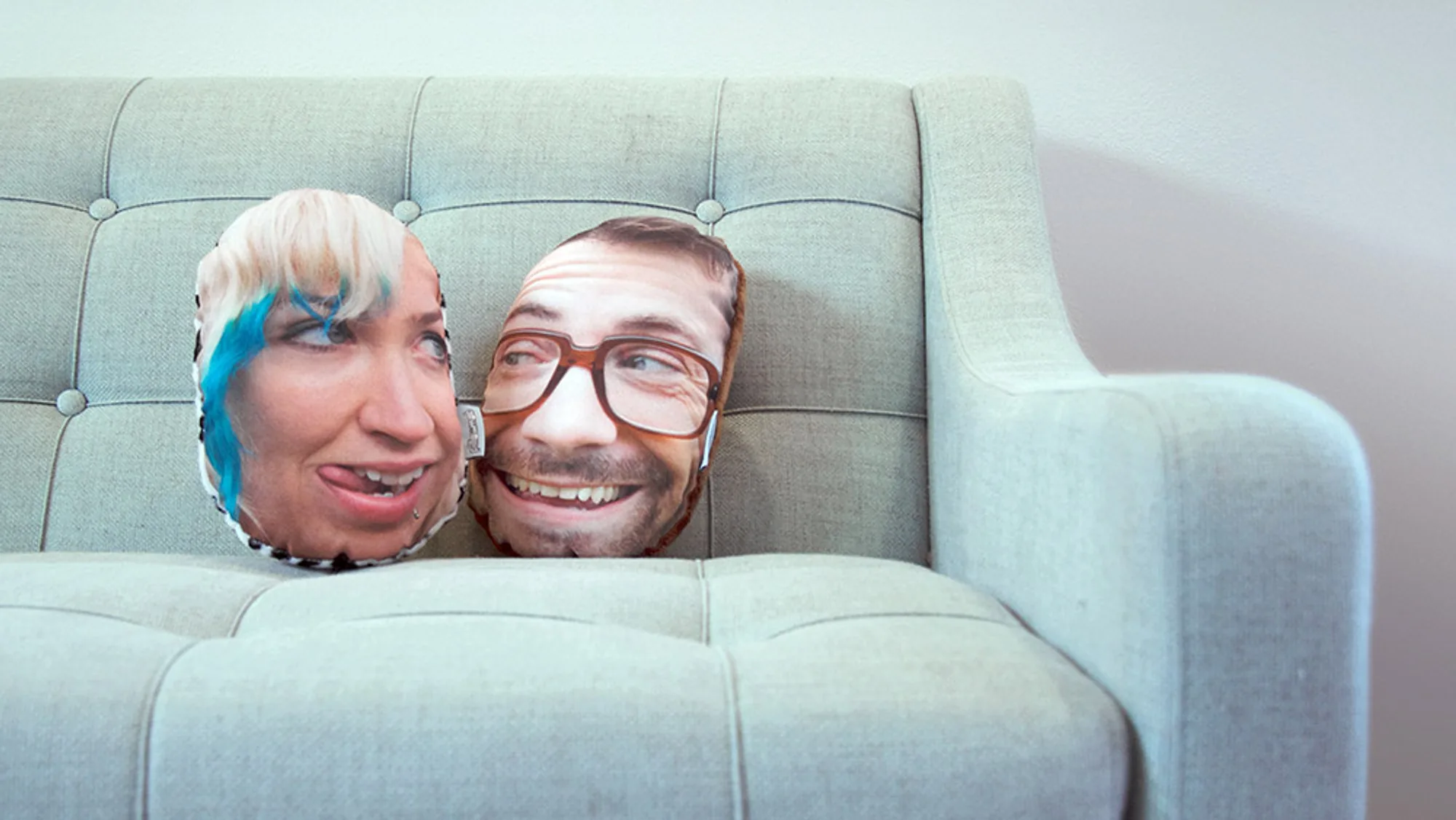 Two face-shaped pillows propped side by side on a pale green sofa: a woman with blue-streaked hair sticking her tongue out, and a grinning man in glasses.